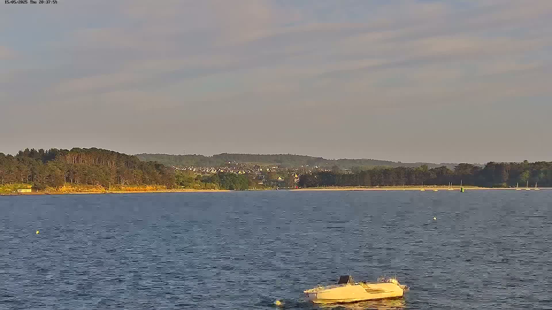 A small white motorboat floats on calm water in front of a wooded shoreline with a town visible in the distance under a partly cloudy sky.