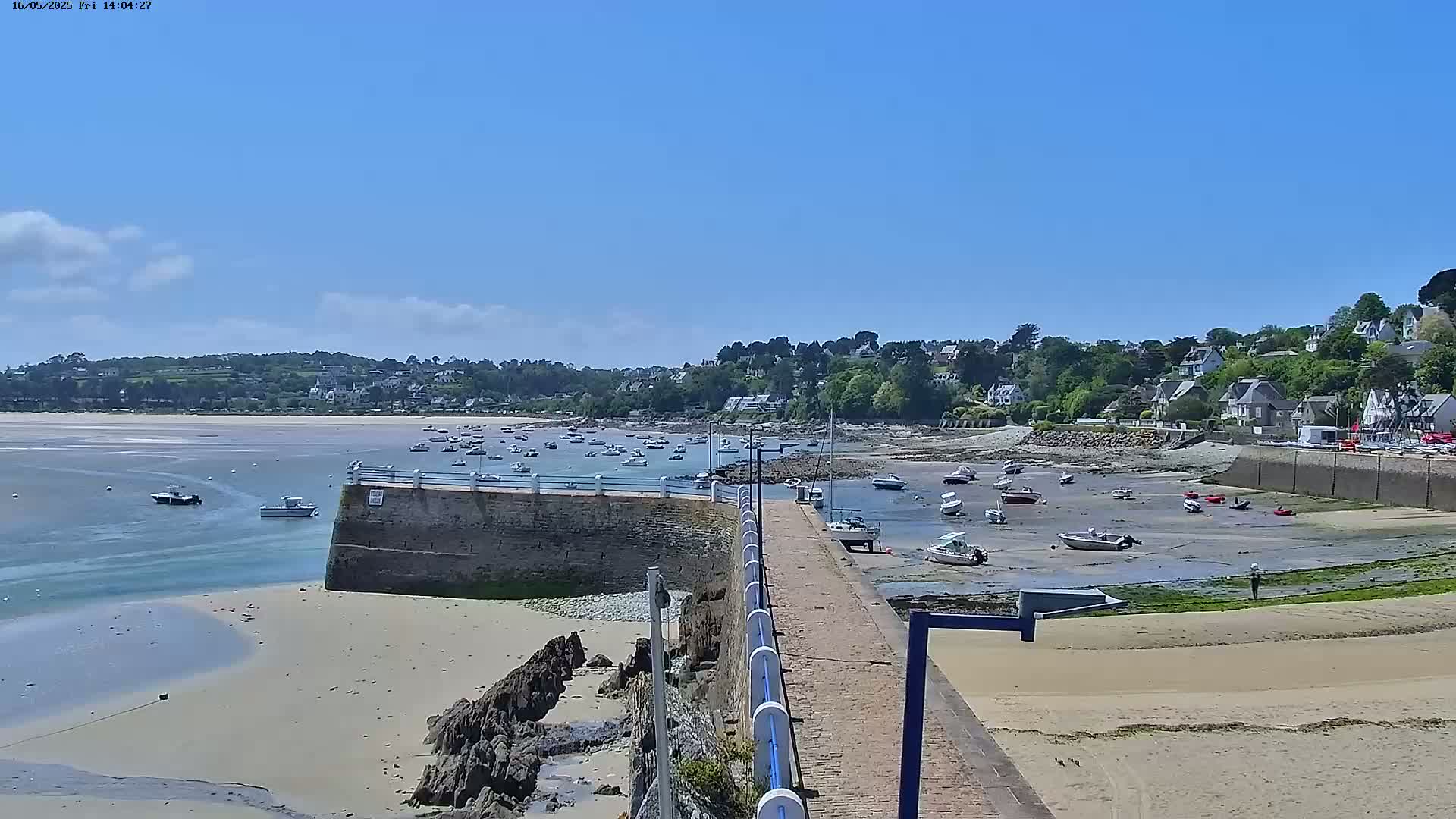 A sunny day reveals a harbor with numerous boats at low tide, a stone pier extending into the water, and a residential area on a hillside in the background.