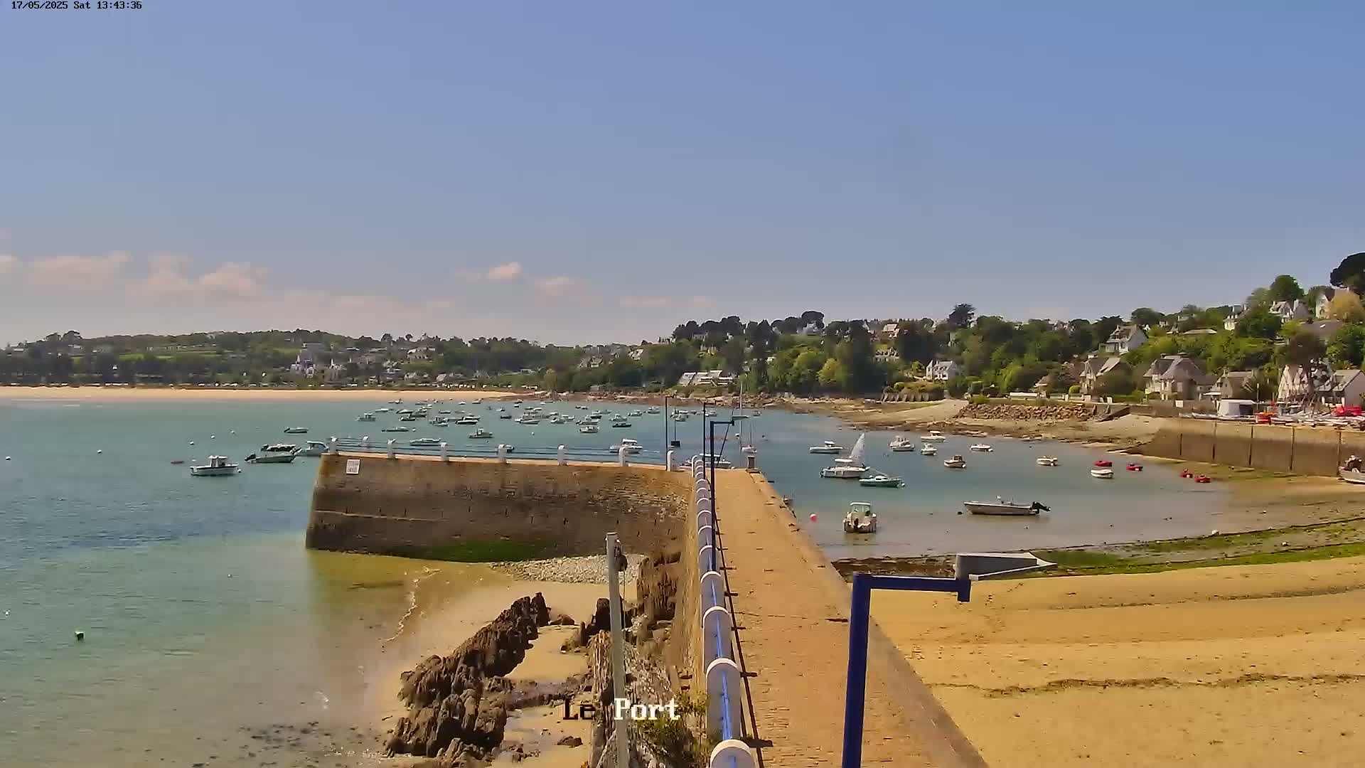 A sunny day reveals a calm harbor filled with numerous boats, a sandy beach, and a stone pier extending into the water, all bordered by a verdant hillside dotted with buildings.