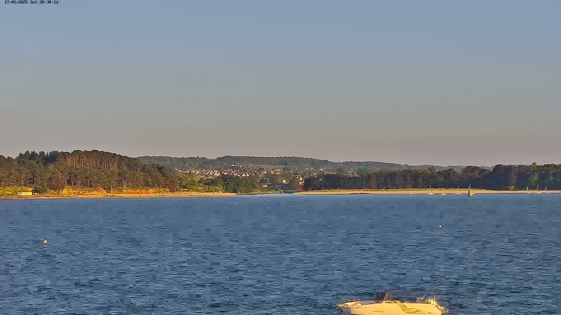 A calm body of water stretches toward a distant shoreline lined with trees and a small town under a clear sky.