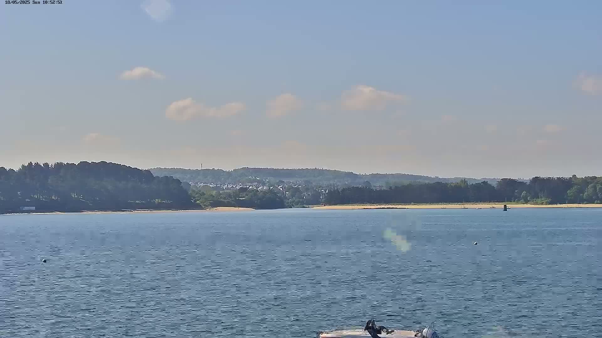 A calm body of water stretches toward a sandy shore lined with trees and a distant town under a mostly clear sky with a few scattered clouds.