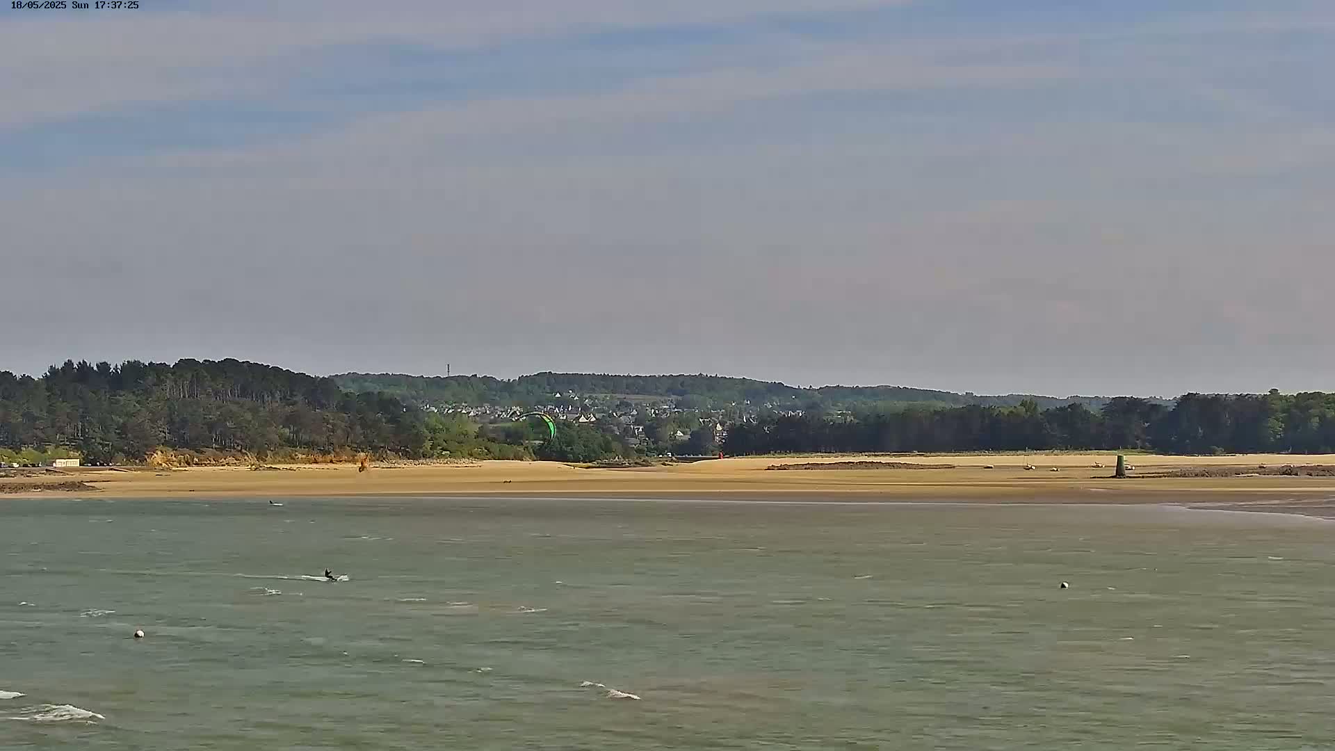 A calm body of water stretches out to a sandy beach backed by a line of trees and a distant town under a mostly clear sky.