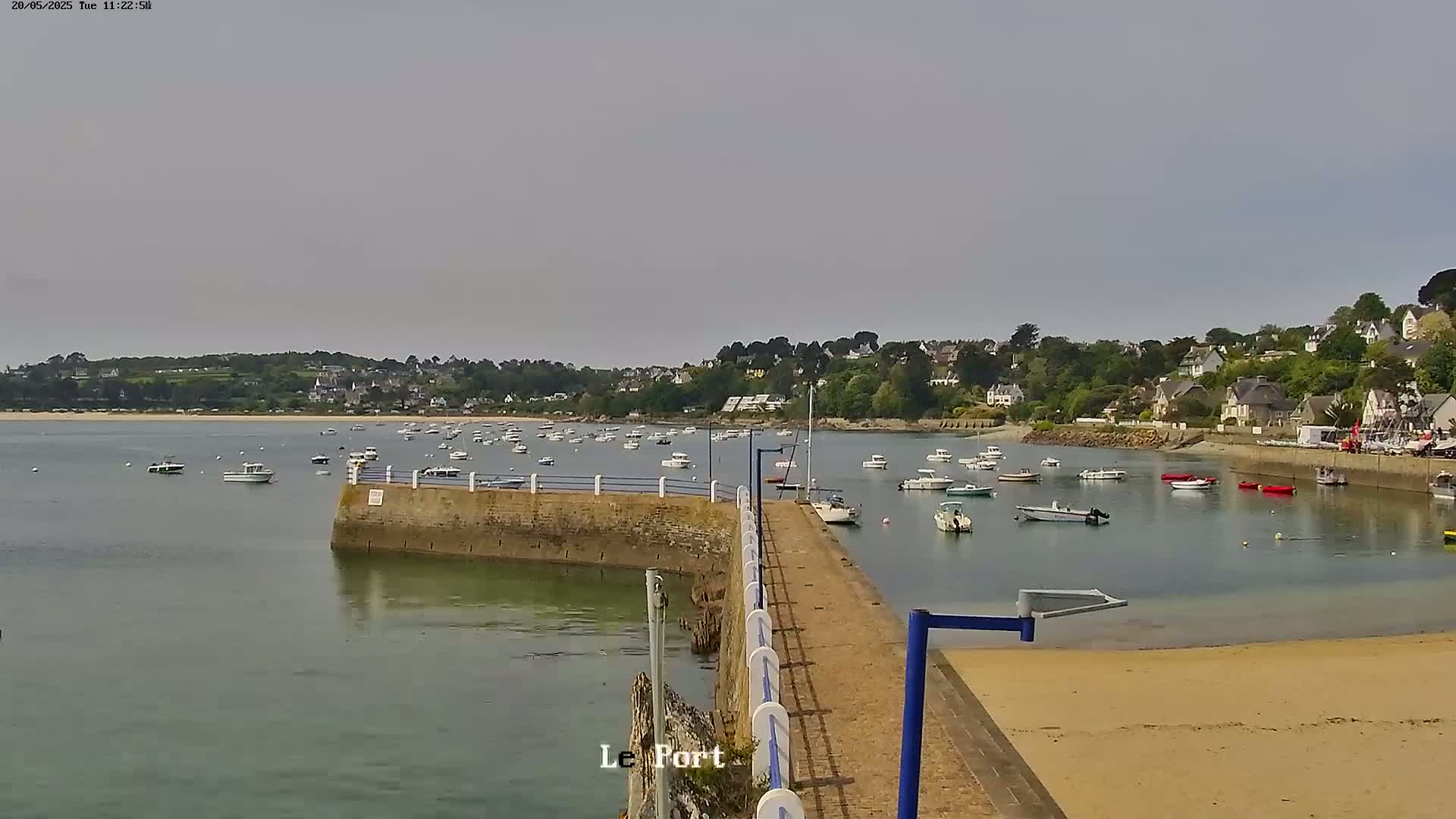 A calm, overcast day shows a harbor filled with numerous small boats, a stone jetty extending into the water, and a sandy beach in the foreground, all backed by a tree-lined hillside with buildings.
