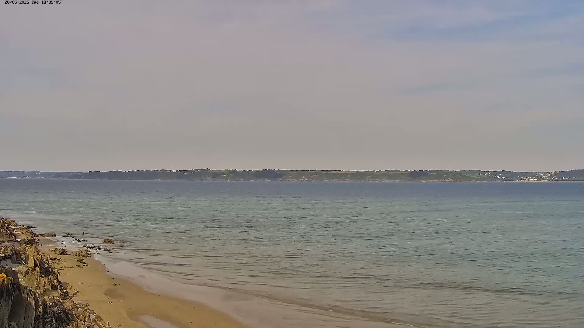 A sandy beach with rocky outcrops meets a calm, light-green sea under a mostly cloudy sky.