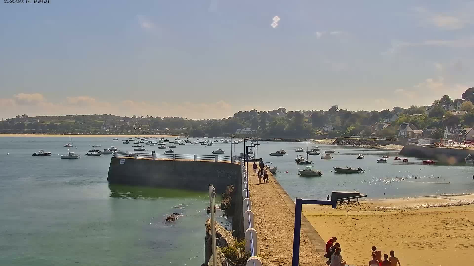 A paved walkway extends from a stone jetty into a calm bay filled with numerous small boats, under a mostly sunny sky with a few scattered clouds, toward a sandy beach and a village nestled amongst green hills.