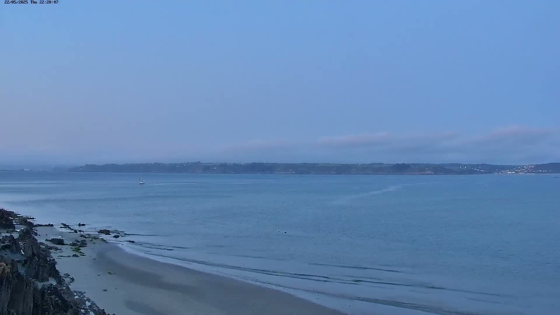 A sandy beach stretches along a rocky shoreline, overlooking a calm body of water with a distant landmass under a clear, twilight sky.
