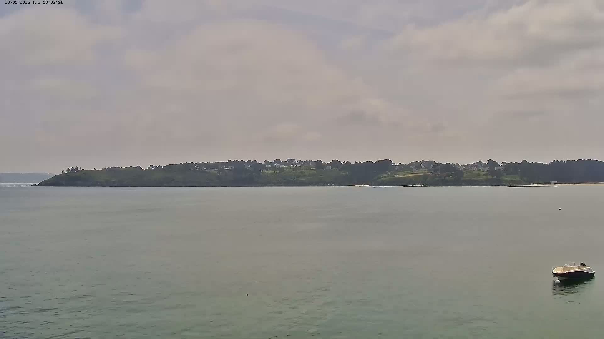 A small boat floats in calm, gray water in front of a distant, tree-covered shoreline under a cloudy sky.