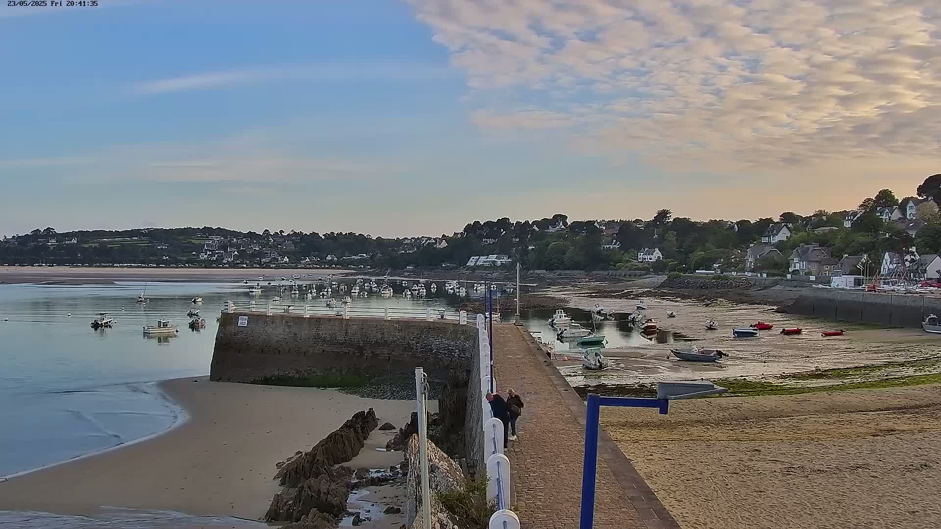 A calm harbor with numerous boats, some grounded on the sand at low tide, is nestled beside a small town under a partly cloudy sky.