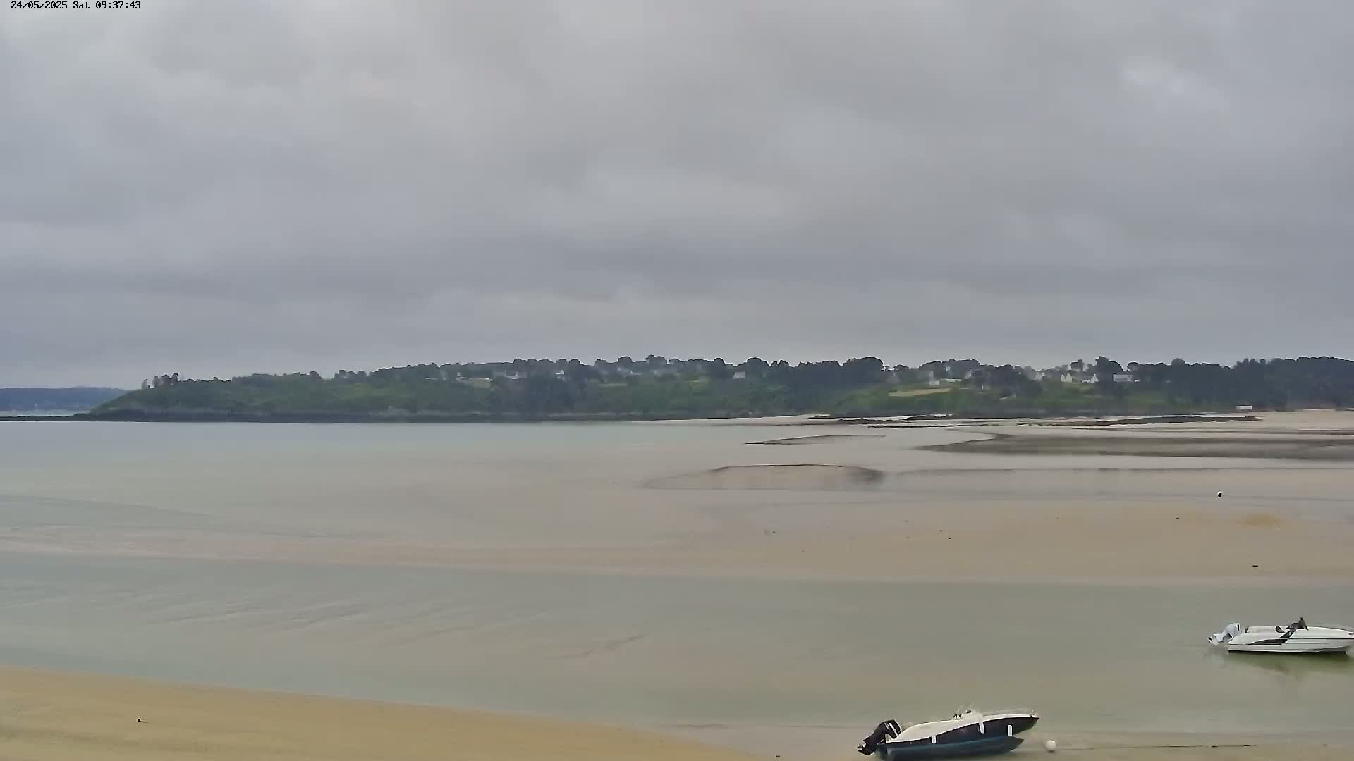 A wide shot of a calm, shallow bay under an overcast sky, showing a sandy shore, two small motorboats, and a tree-covered hill in the distance.