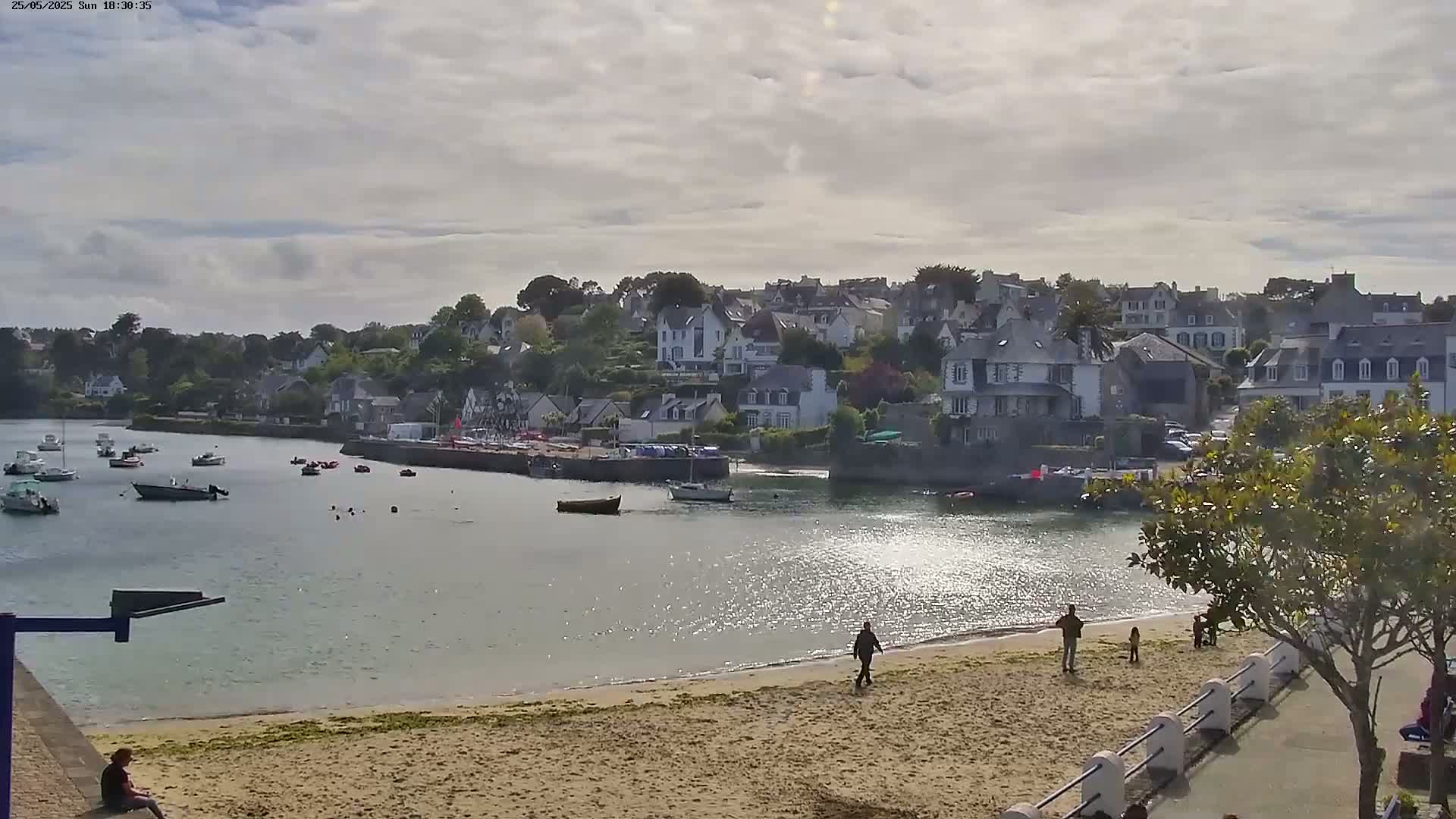 A sandy beach with a few people on it fronts a calm bay with several boats, overlooked by a village of closely-packed houses under a partly cloudy sky.