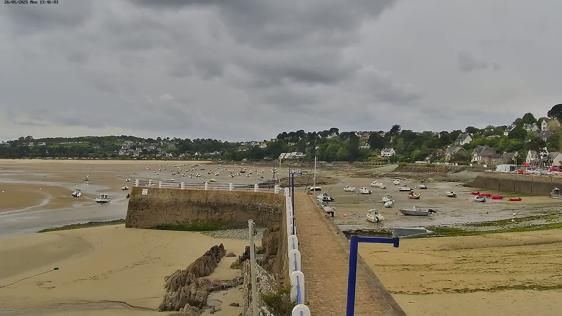 A sandy beach with numerous small boats resting on the exposed seabed during low tide under an overcast sky, backed by a village nestled amongst green hills.