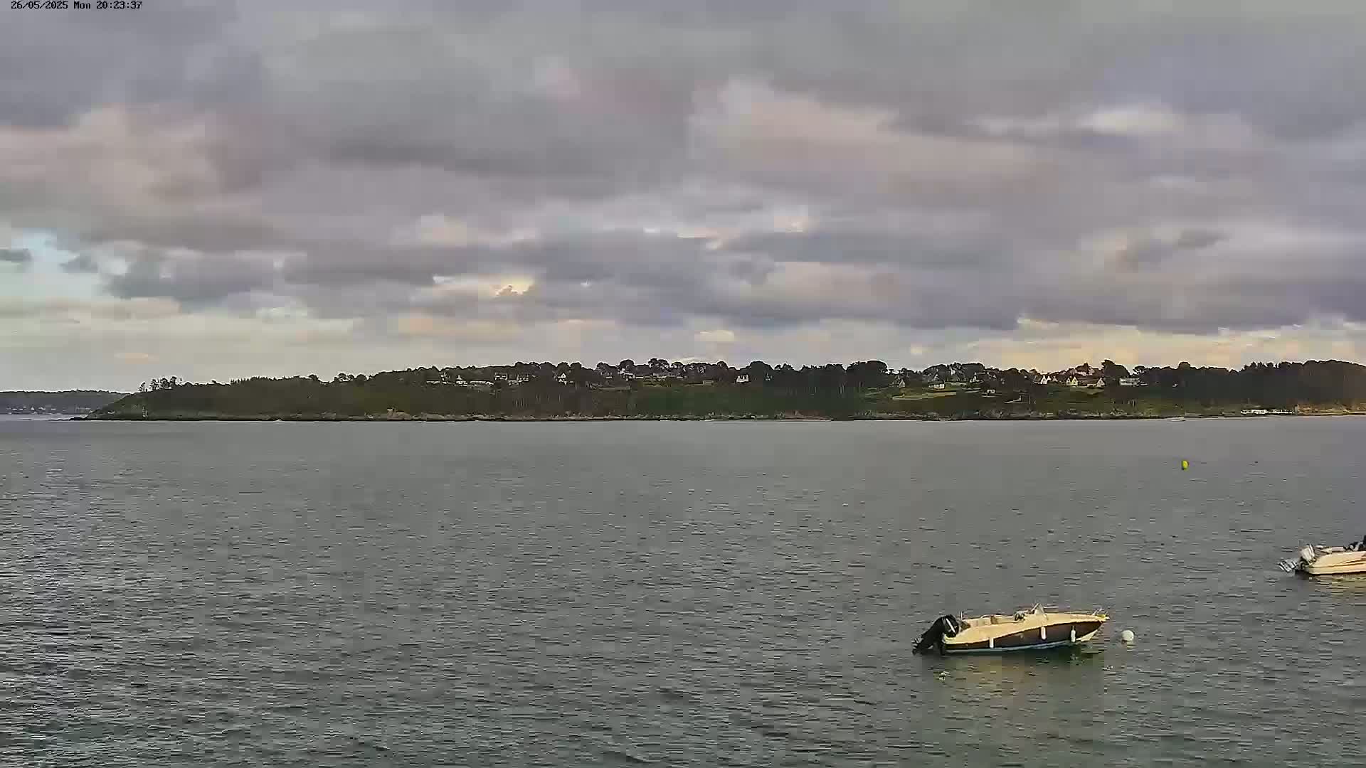 Two small motorboats are anchored in calm water in front of a tree-covered shoreline under a mostly cloudy sky.