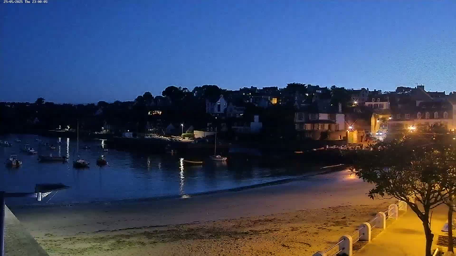 A sandy beach at night is illuminated by streetlights, overlooking a calm harbor filled with boats and a village of buildings on a hill under a dark blue twilight sky.
