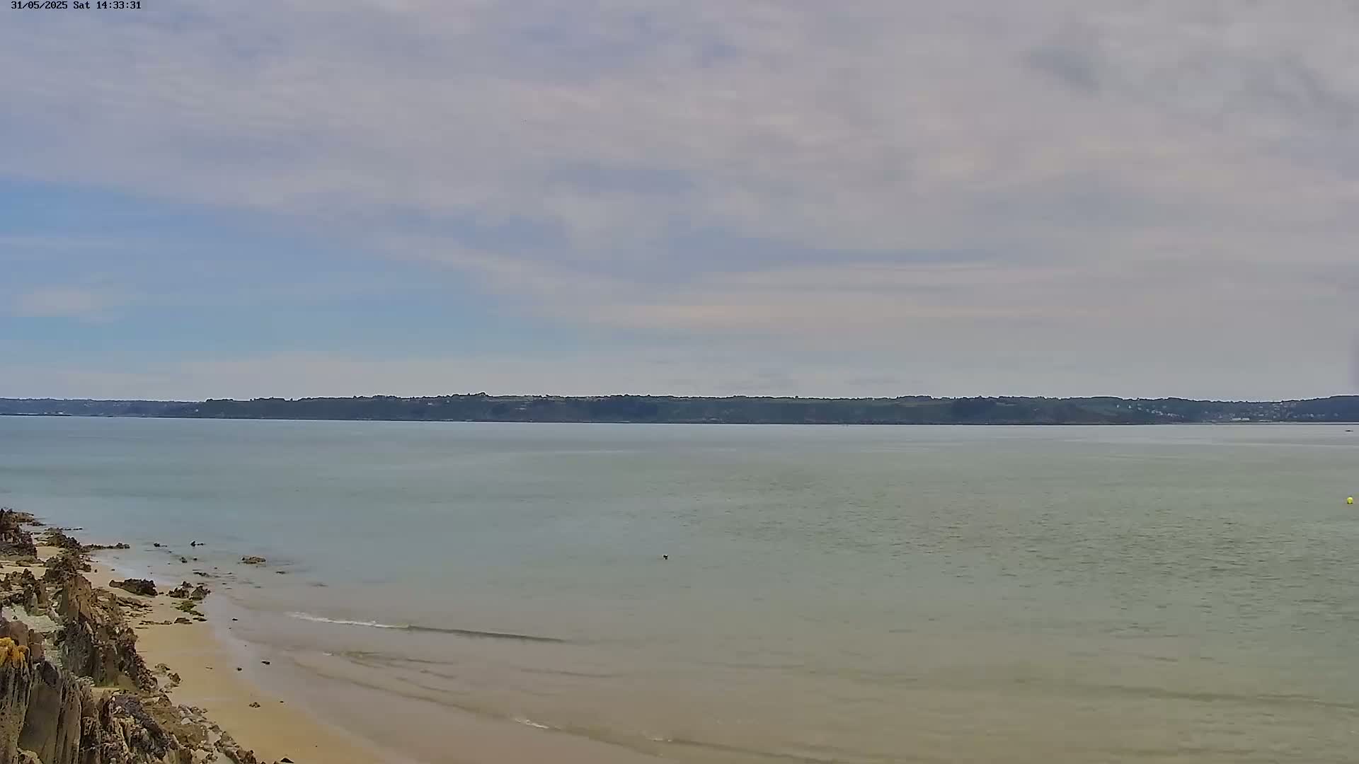 A calm, light-grey sea stretches to a distant, dark green shoreline under a mostly cloudy sky.