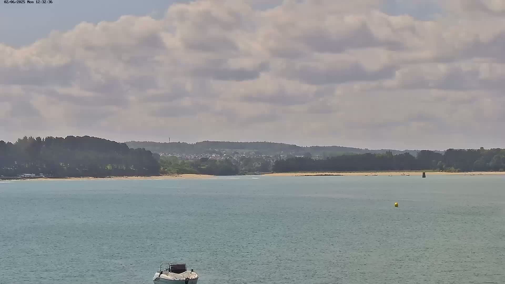 A partly cloudy day shows a calm body of water with a small boat in the foreground, a sandy beach and dark green trees in the middle ground, and a town in the distant background.