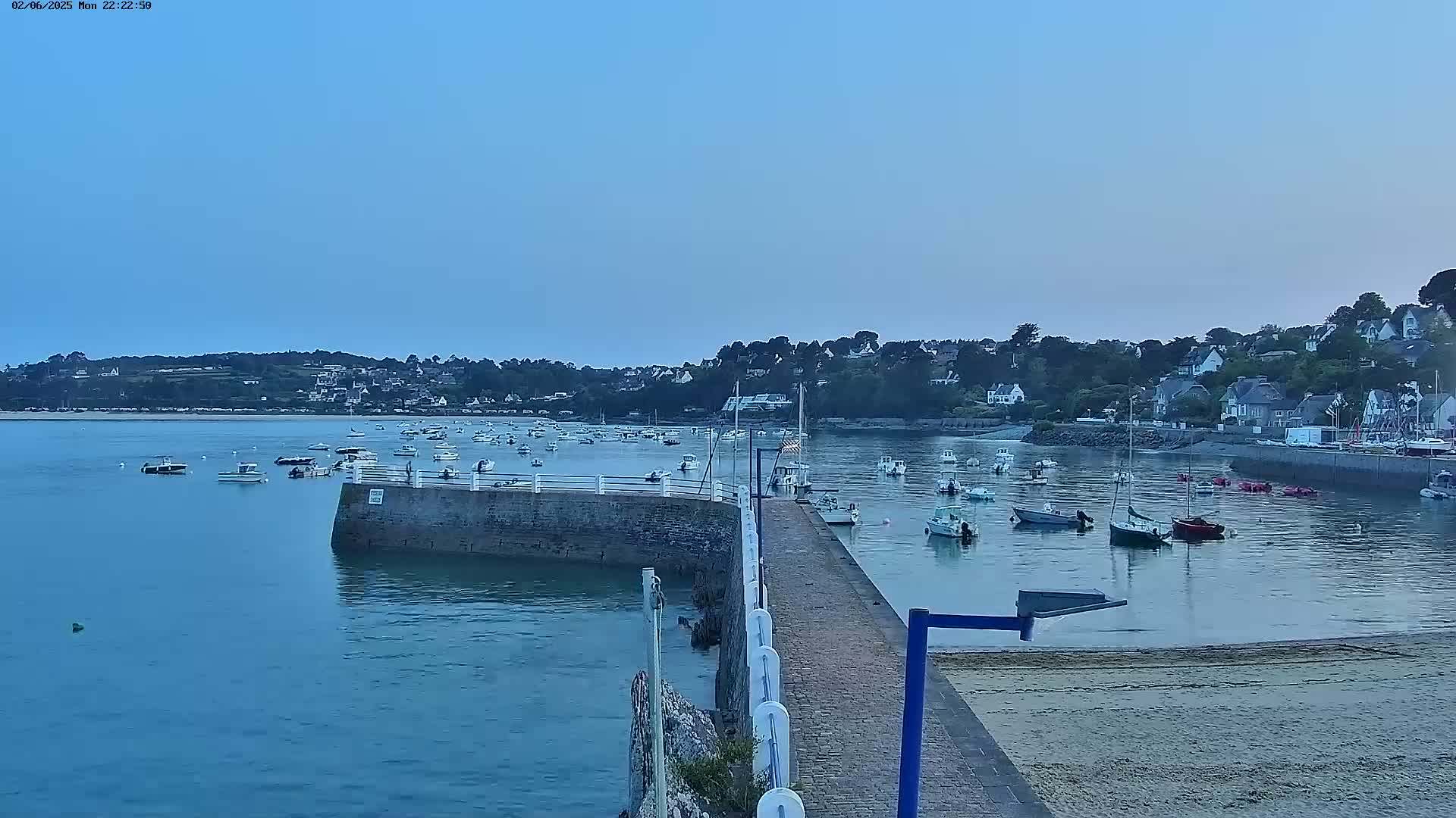 A calm, twilight harbor scene shows numerous small boats moored near a stone pier and a village nestled on a hillside in the background.