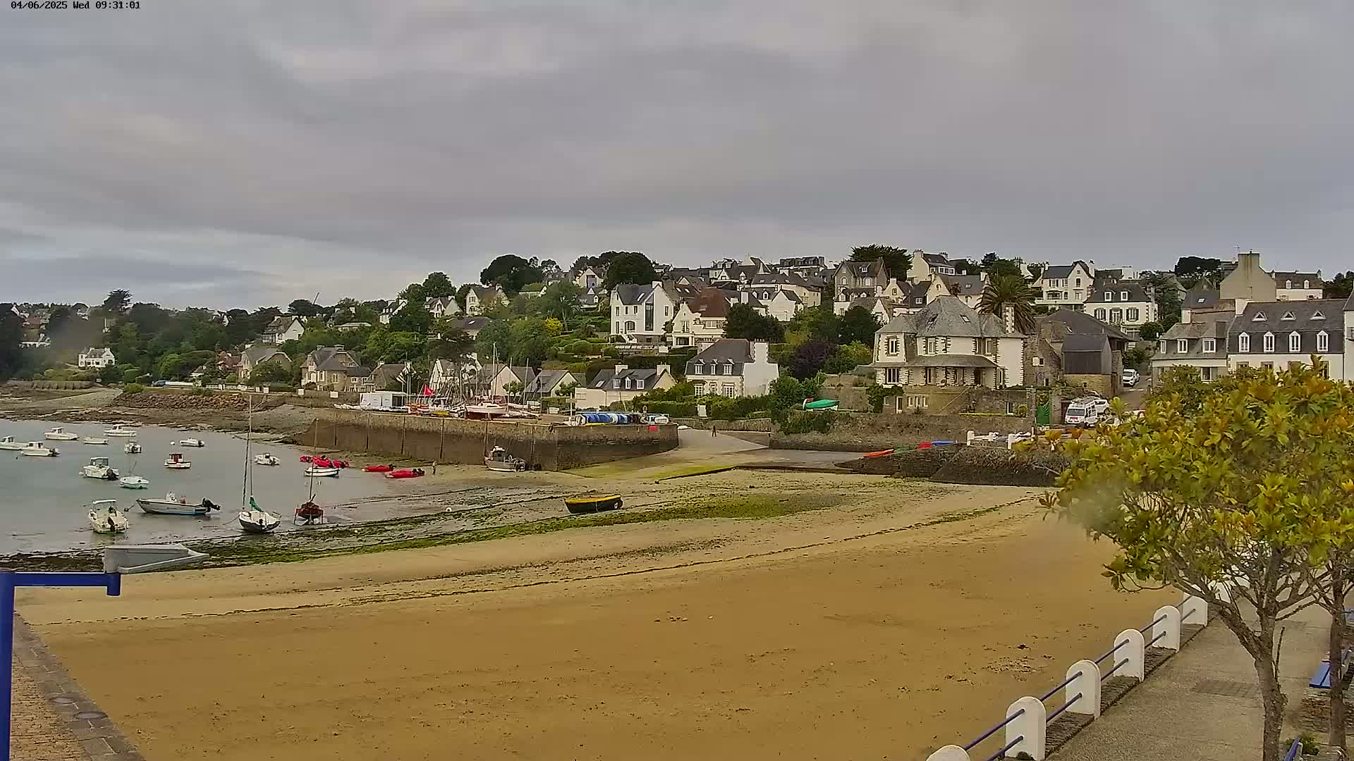 A sandy beach stretches out before a small coastal town under a cloudy sky, with numerous boats moored in the shallow water at low tide.