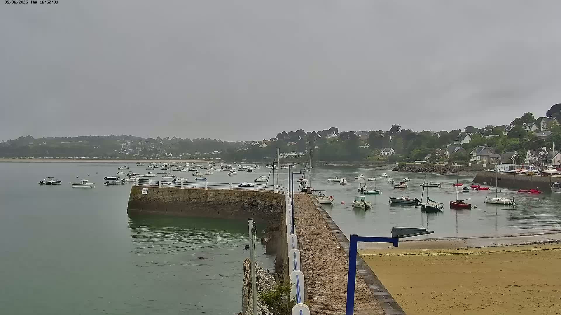 A harbor filled with numerous boats is seen under an overcast sky, with a stone walkway and sandy beach in the foreground.