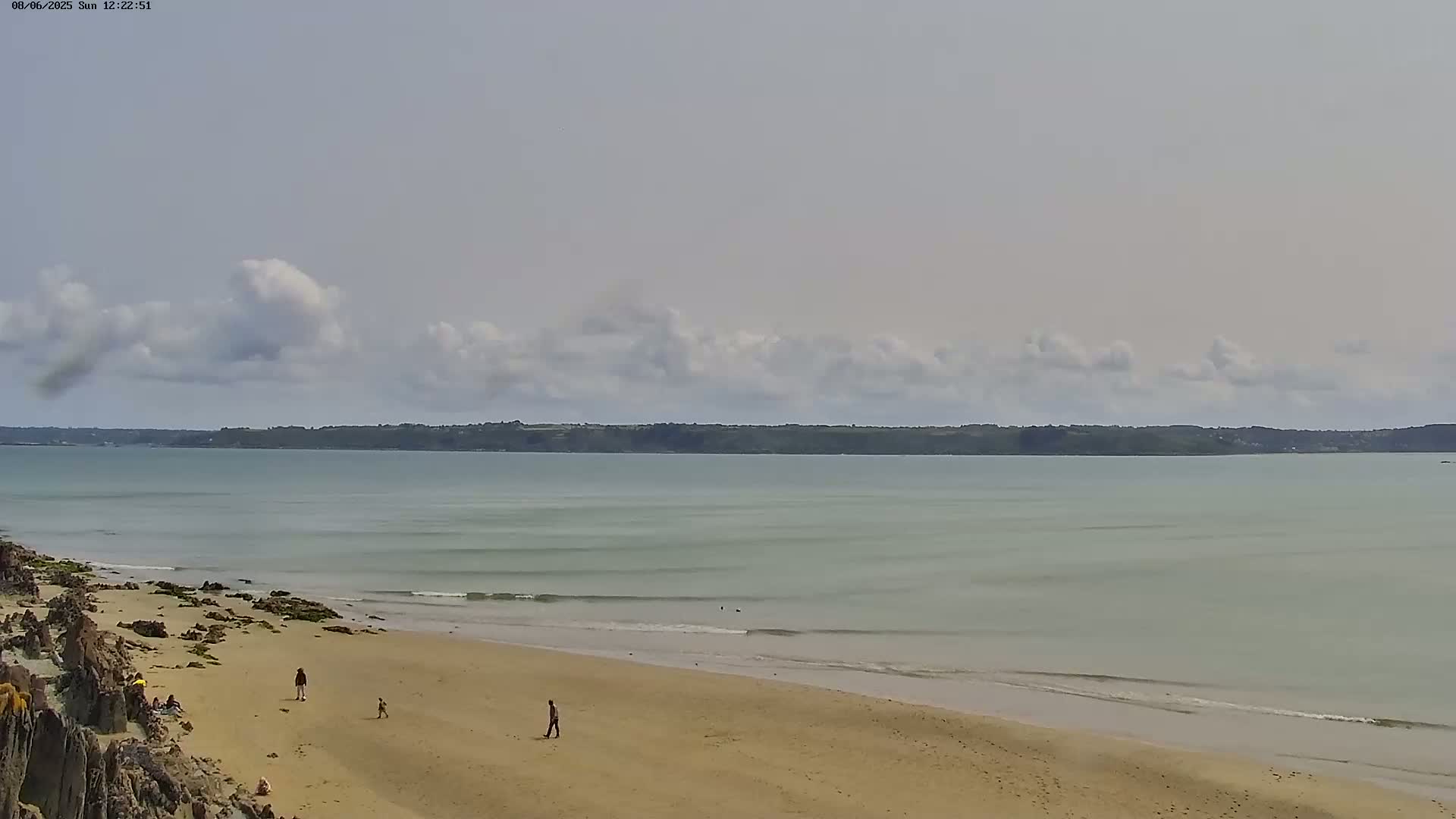 A sandy beach with a few people on it stretches out to meet a calm, light-green sea under a partly cloudy sky.