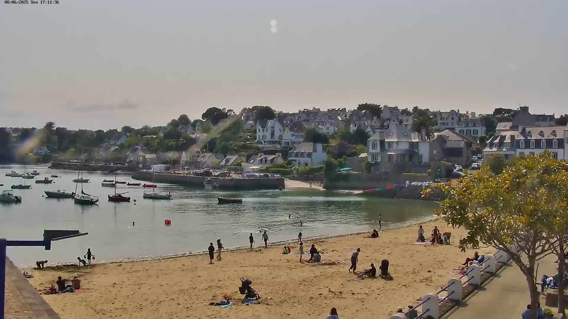 A sandy beach with scattered people on a sunny day overlooks a calm harbor filled with boats, and a small town is visible beyond the harbor.