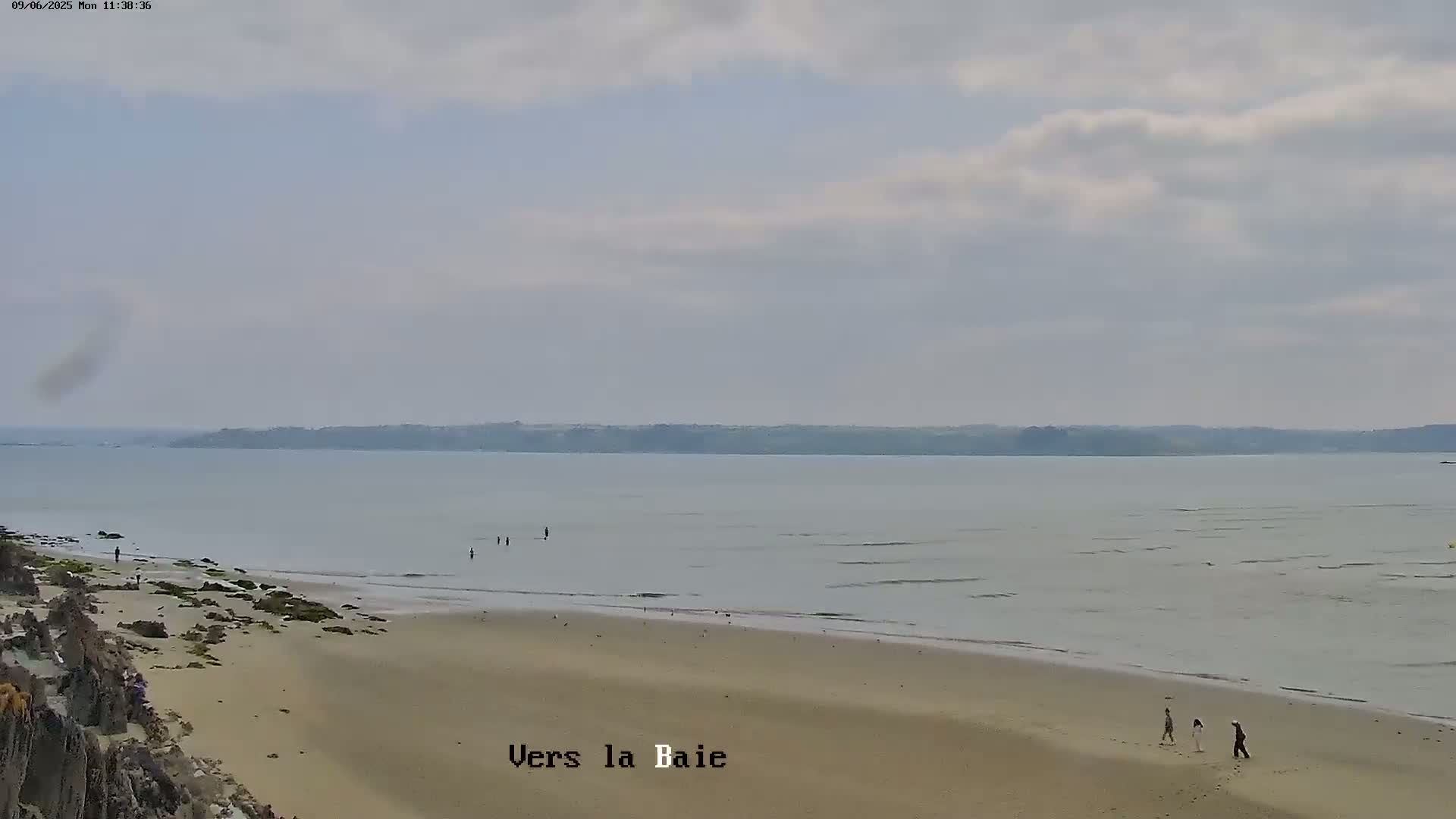 A sandy beach stretches toward a calm, overcast ocean with a few people walking along the shore and others in the water.