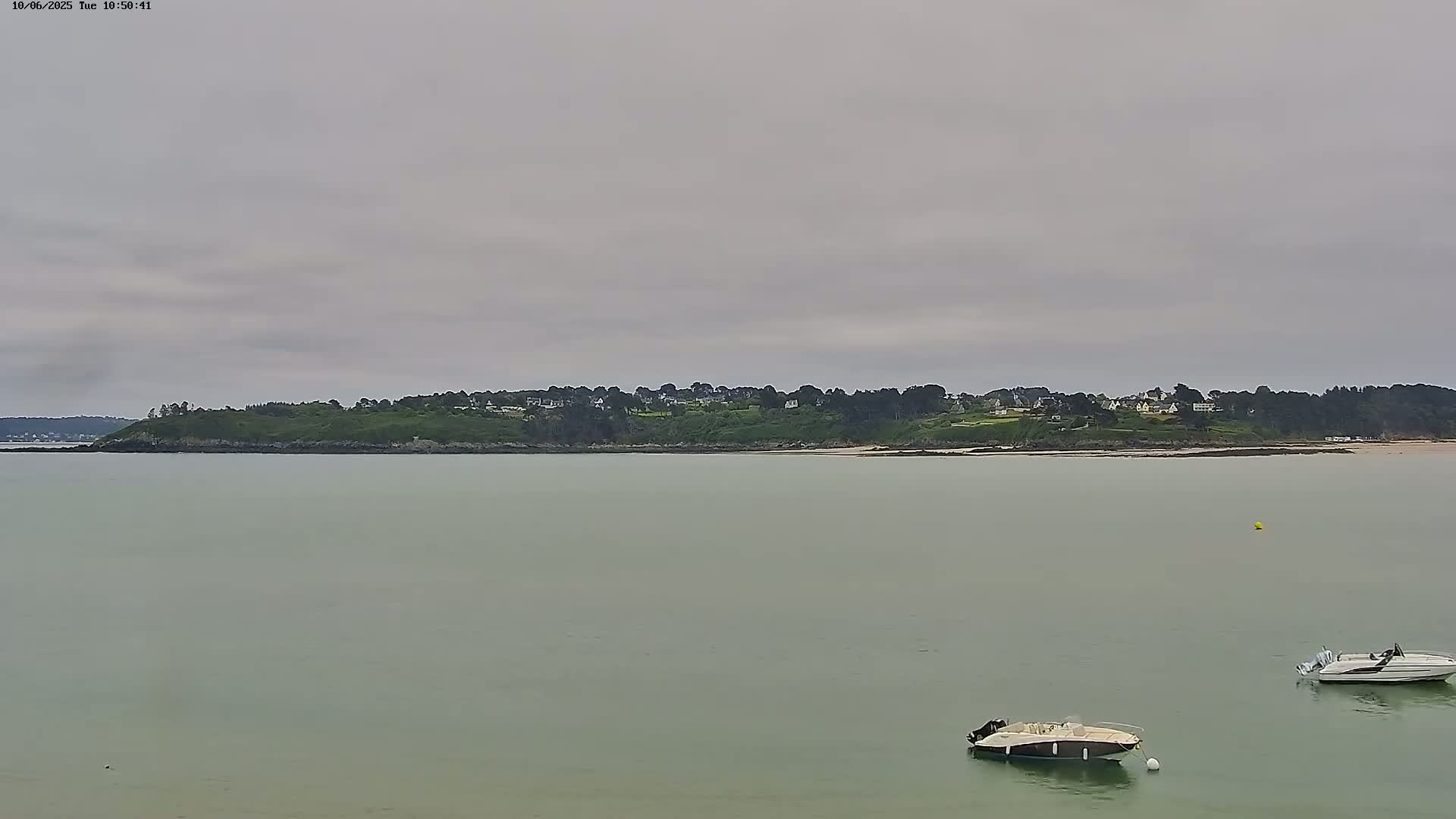A calm, overcast day shows a body of water with two small motorboats and a distant shoreline featuring a line of green hills and houses.