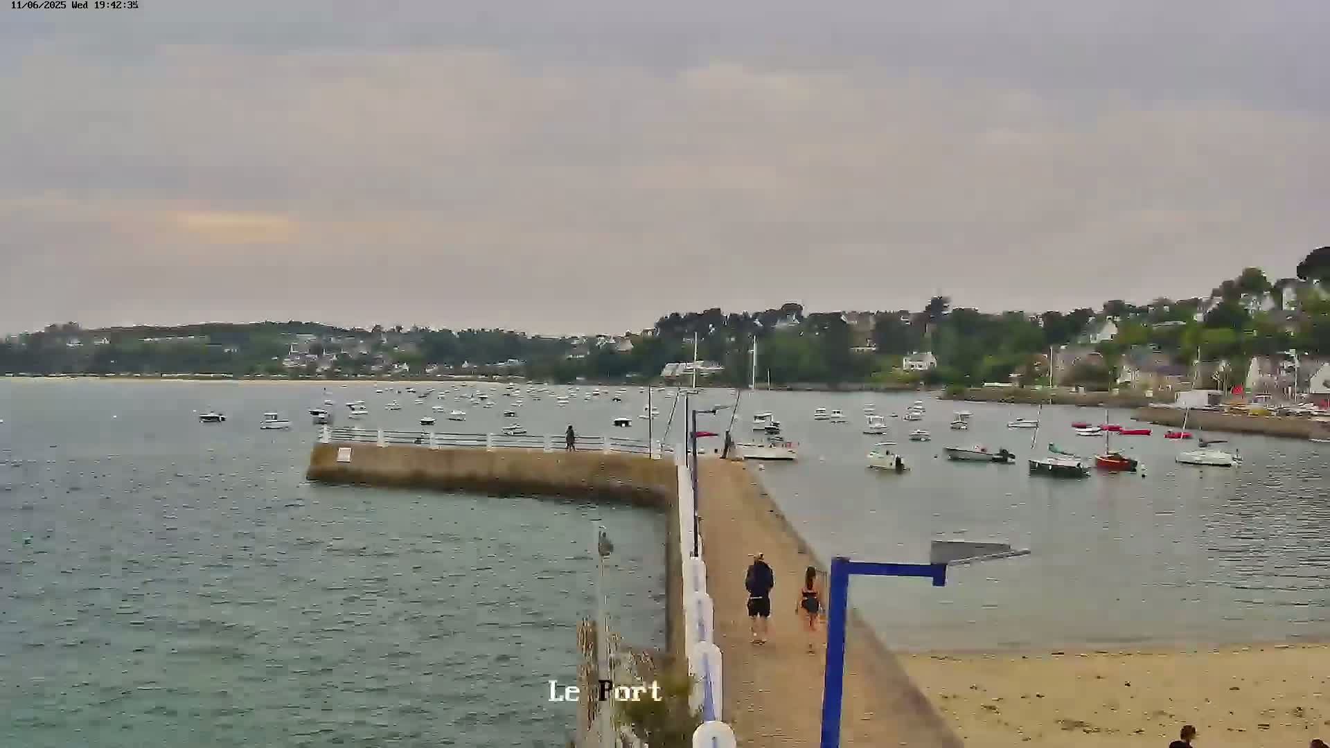 A harbor filled with numerous boats is viewed from a pier on an overcast day, with a few people walking along the pier.