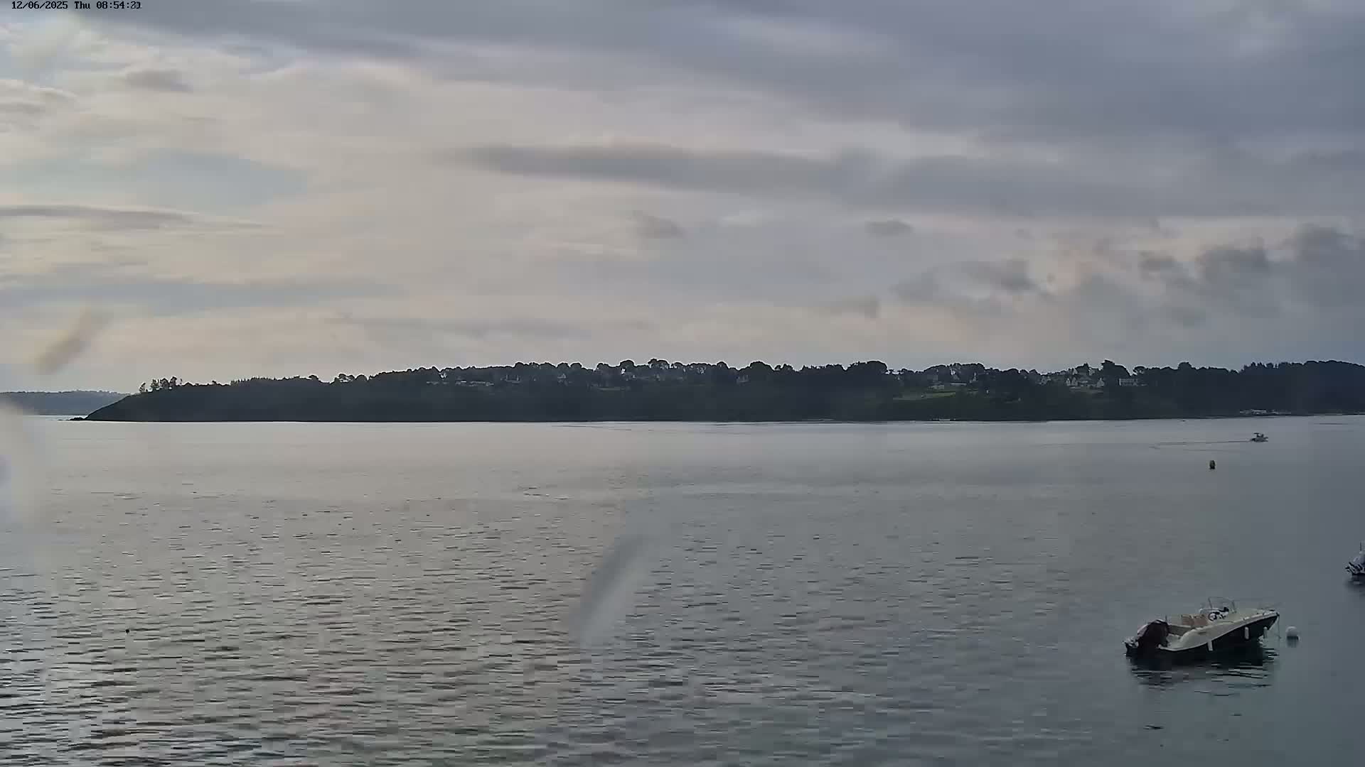 A calm body of water with a few boats is seen in front of a tree-lined shoreline under an overcast sky.