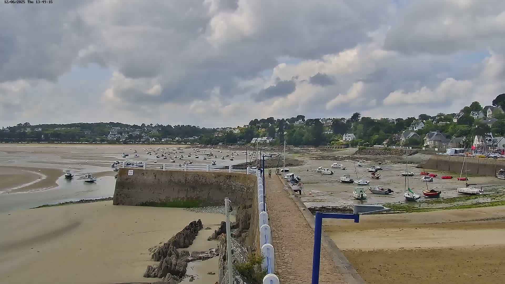 A sandy harbor with numerous boats at low tide, under a partly cloudy sky, is overlooked by a village on a hillside.