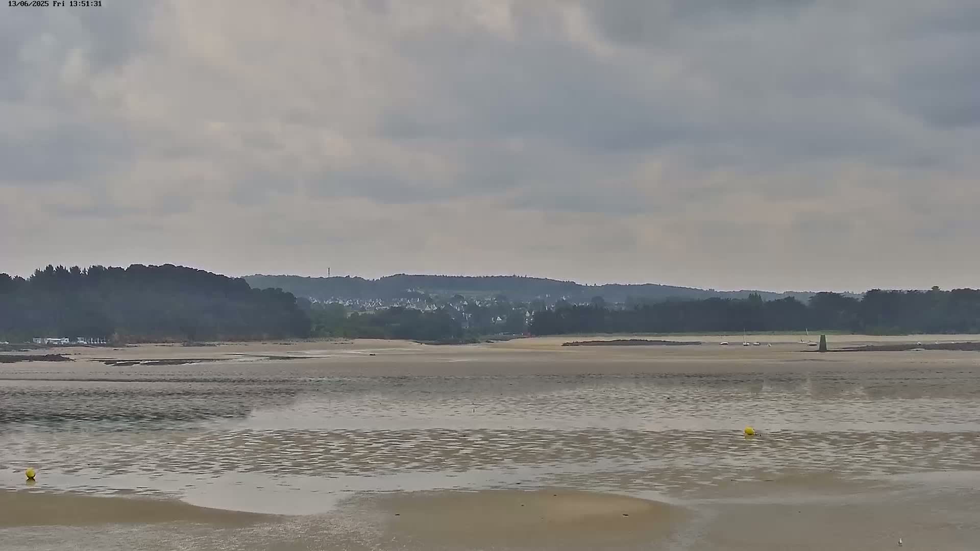 A wide expanse of shallow, rippled water stretches across a sandy bay towards a distant line of trees and buildings under a cloudy sky.