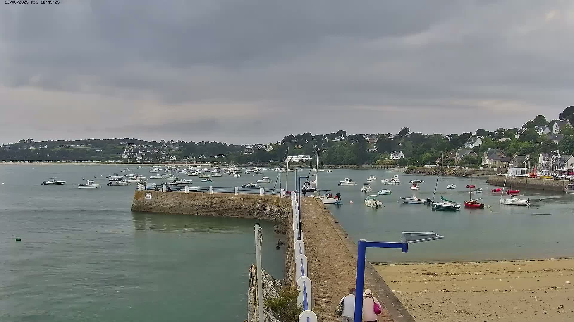 A stone pier extends into a calm harbor filled with numerous small boats under an overcast sky, with a sandy beach and a village visible in the background.