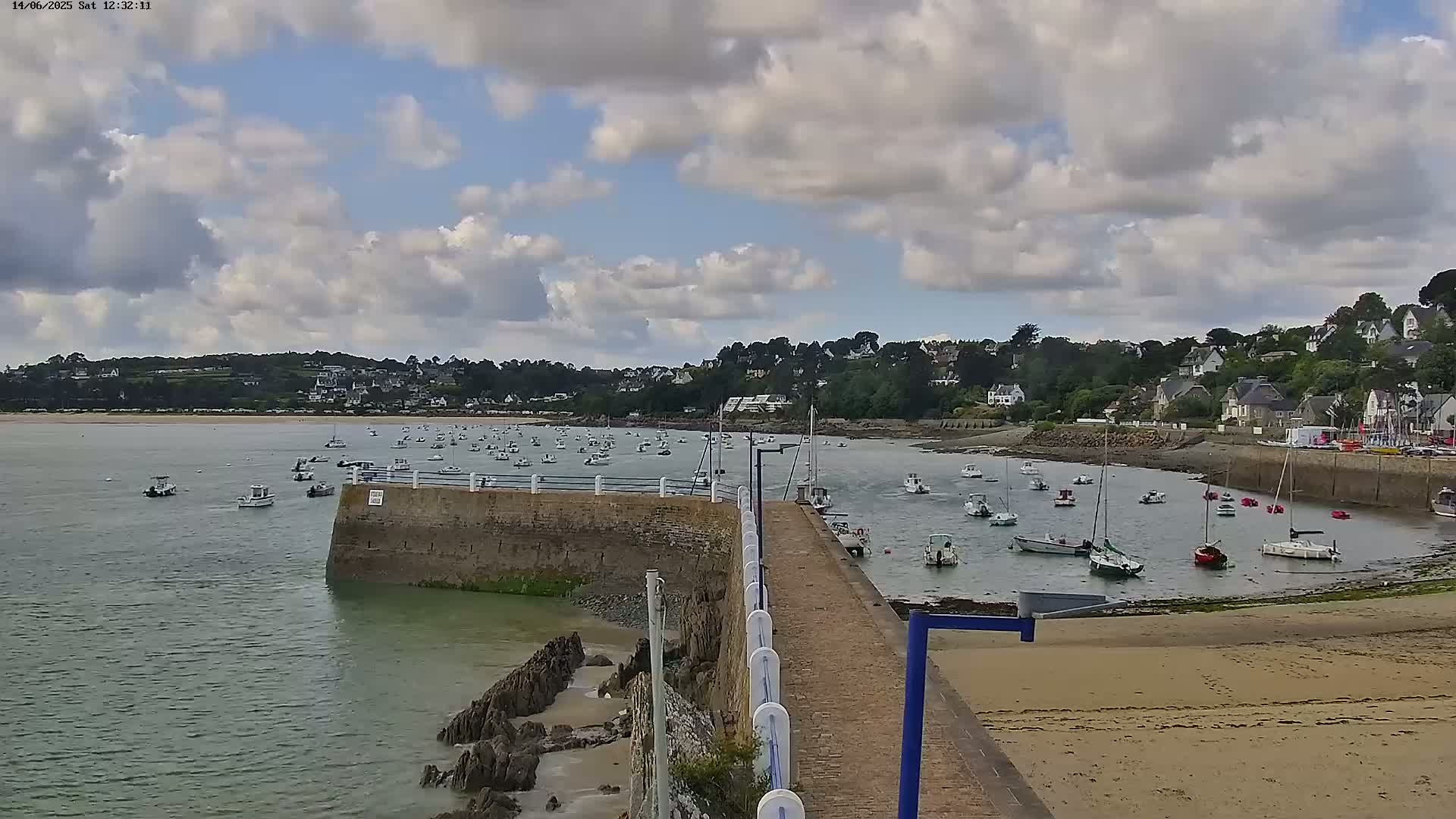 A partly cloudy day reveals a calm harbor filled with numerous boats, nestled beside a small sandy beach and a hillside village.