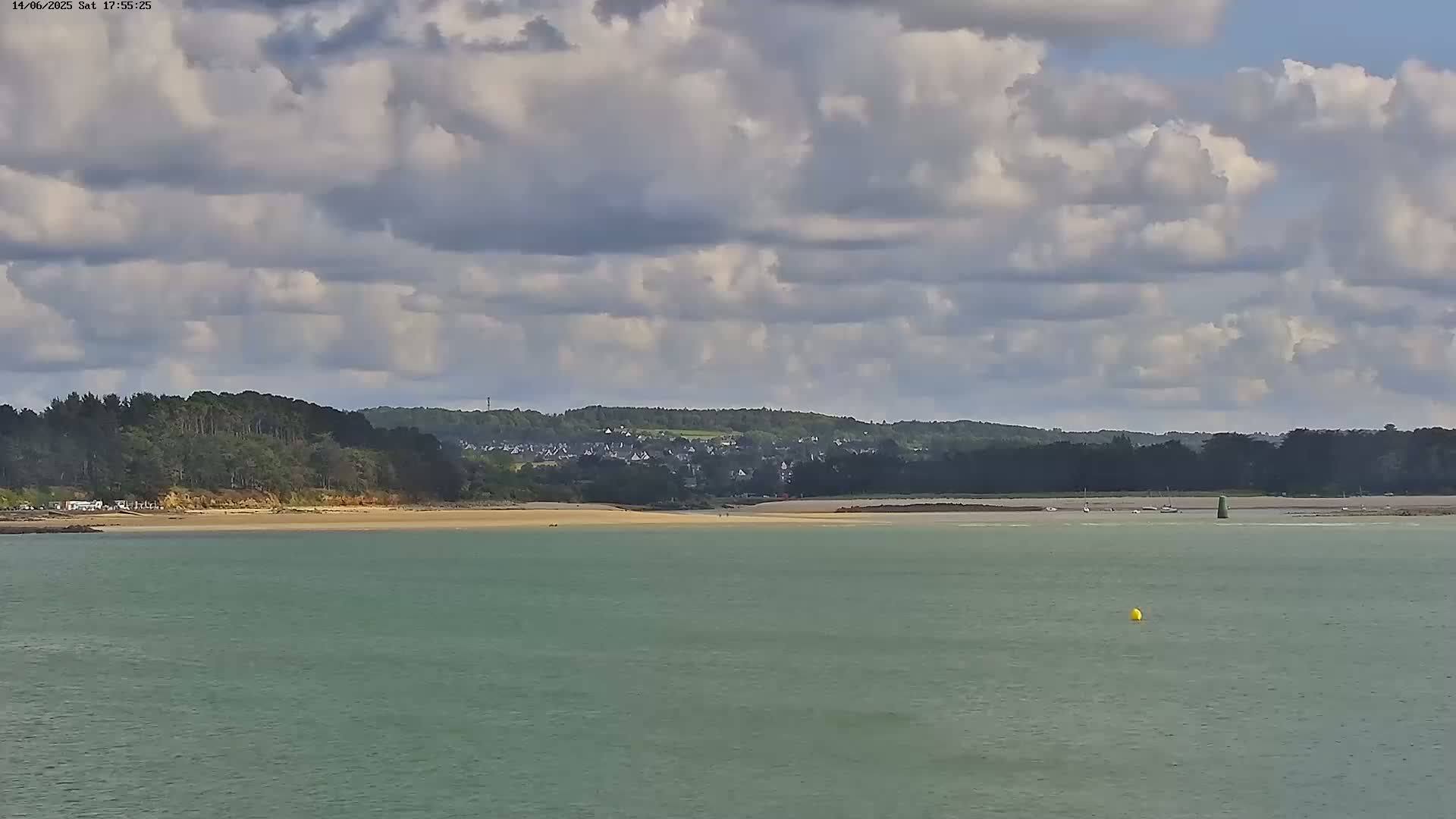 A calm body of water stretches towards a sandy beach backed by a line of trees and a distant village under a partly cloudy sky.