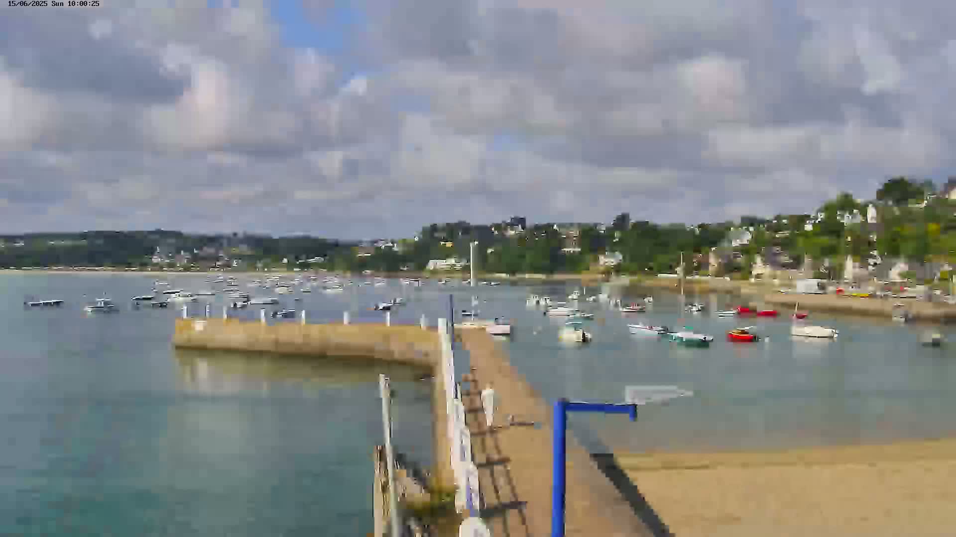 A partly cloudy day shows a calm harbor filled with numerous small boats near a shoreline with buildings and trees.
