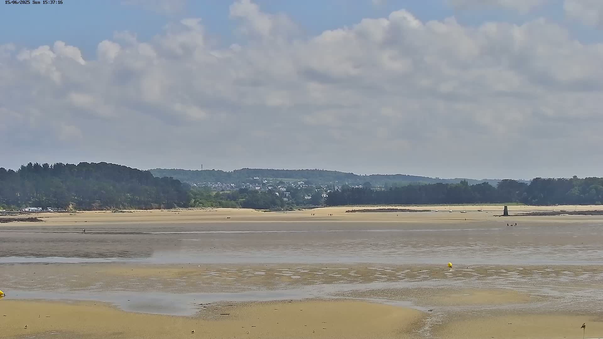 A wide expanse of sand and shallow water at low tide stretches to a distant shoreline of trees and buildings under a partly cloudy sky.