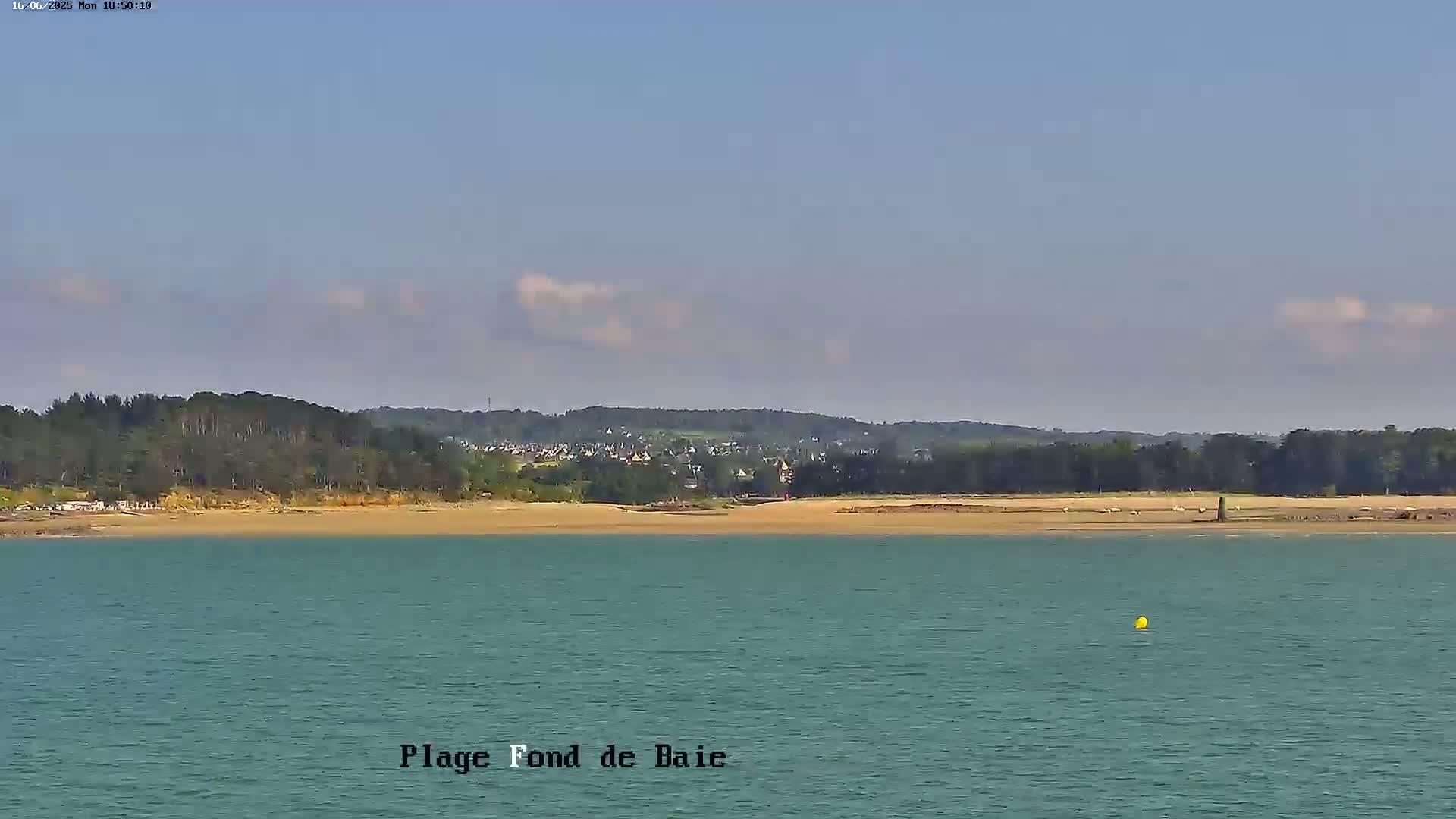 A sandy beach backed by trees and a small town is visible across a calm body of water under a mostly clear sky with a few scattered clouds.