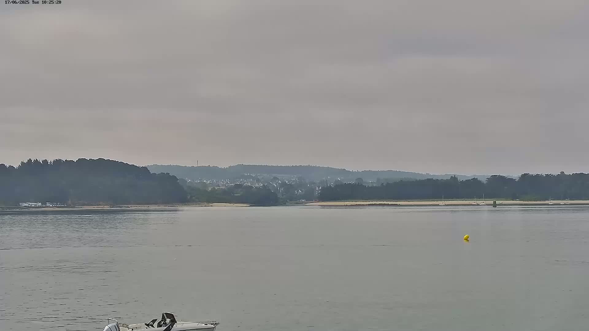A calm, overcast day shows a body of water with a small boat in the foreground and a distant shoreline with trees and buildings under a gray sky.
