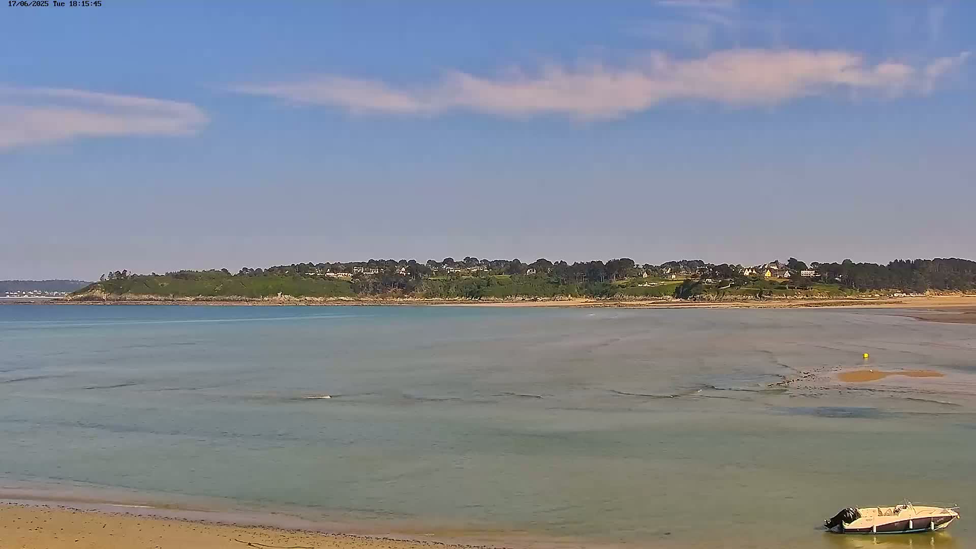A tranquil, shallow body of water stretches toward a verdant headland under a mostly clear, sunny sky, with a small boat beached near the sandy shore in the foreground.