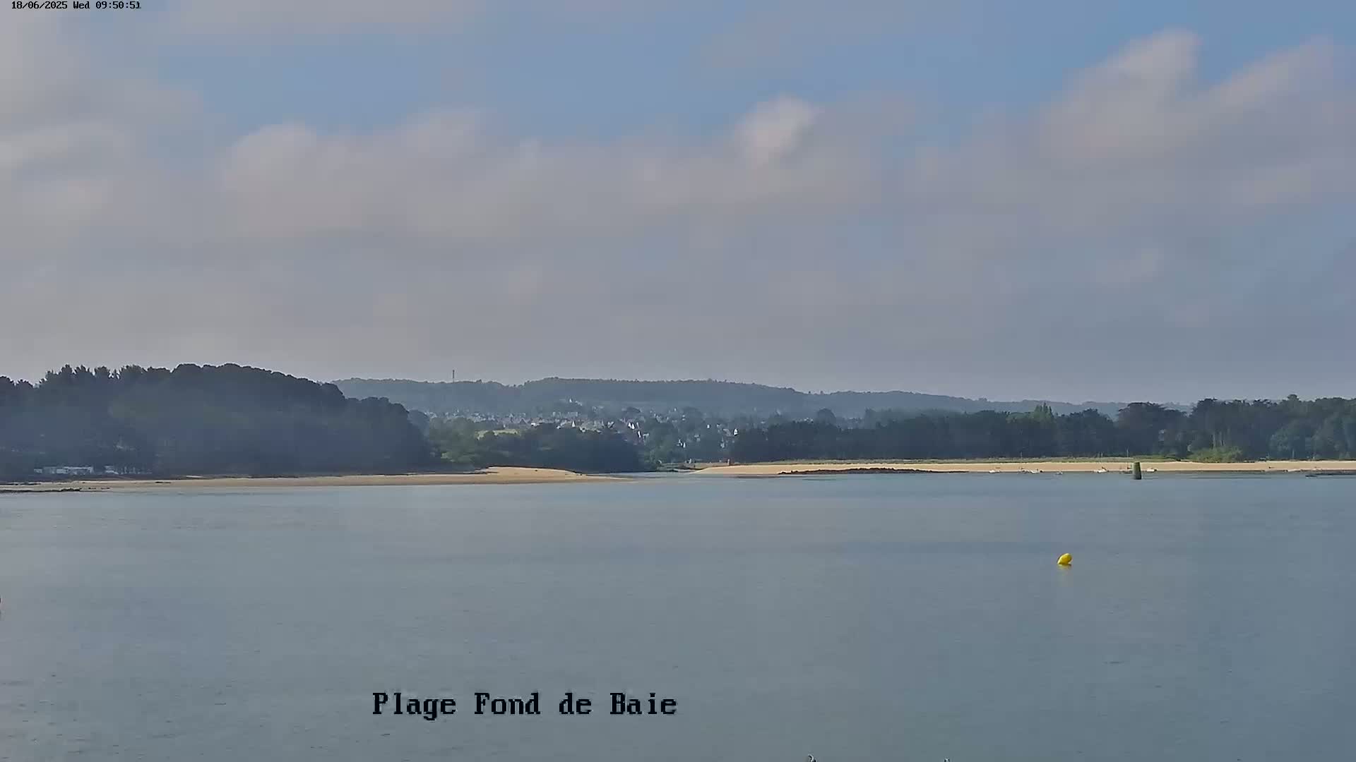 A calm body of water stretches out to a sandy beach backed by a line of trees and a distant town under a partly cloudy sky.