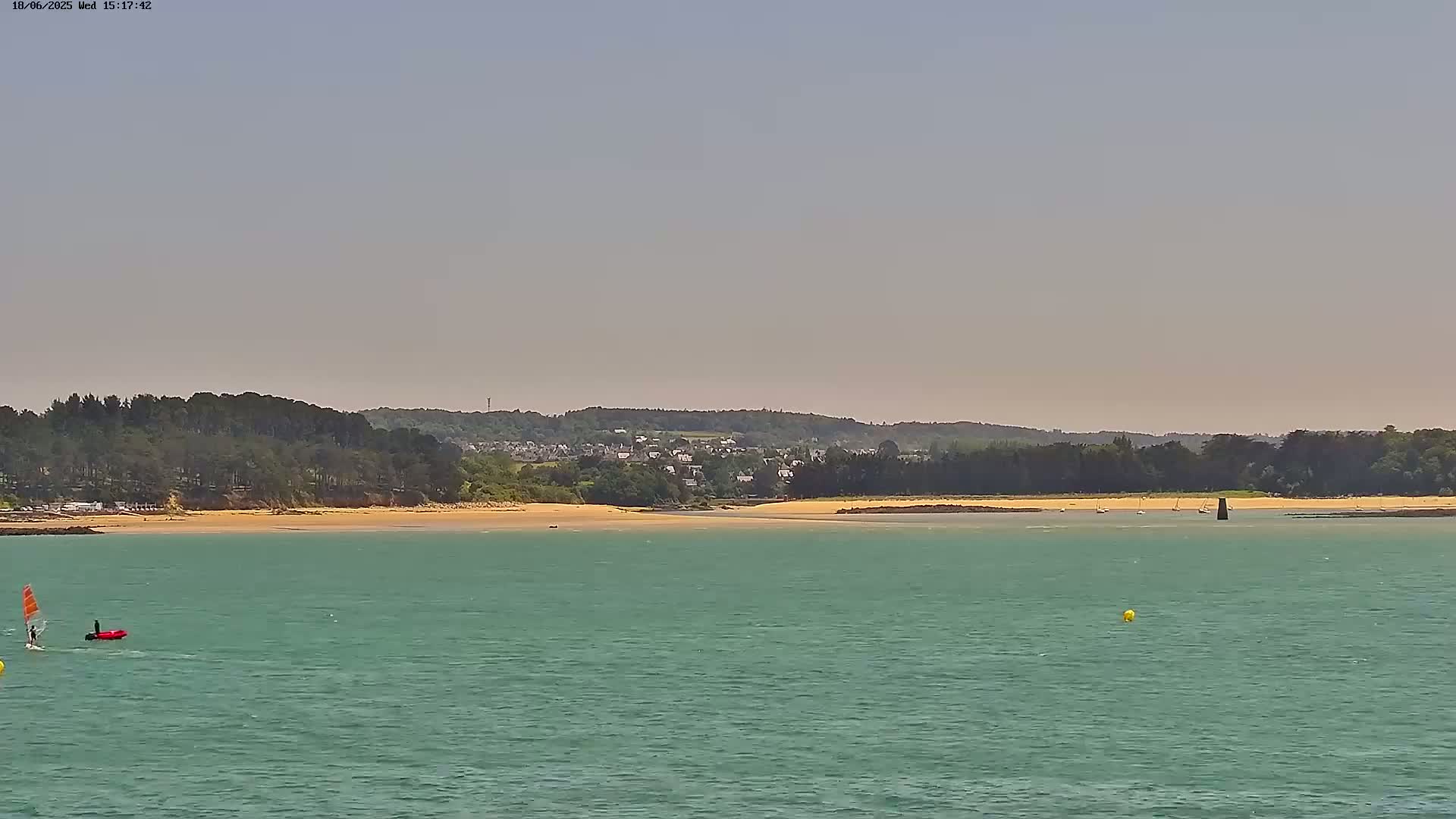 A windsurfer and small boat are visible on calm, teal water in front of a sandy beach and a line of trees under a hazy sky.