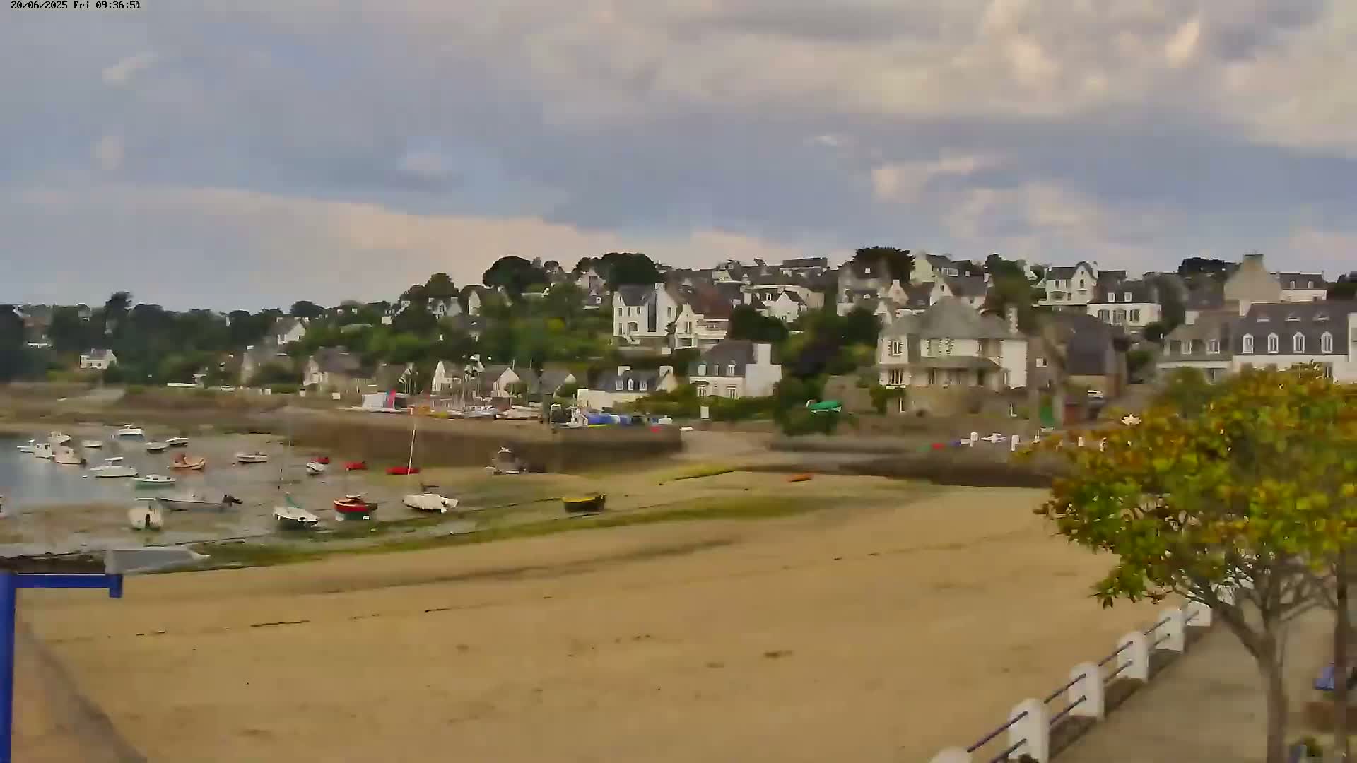 A sandy beach with numerous small boats rests in the shallows, overlooking a hillside village under a partly cloudy sky.
