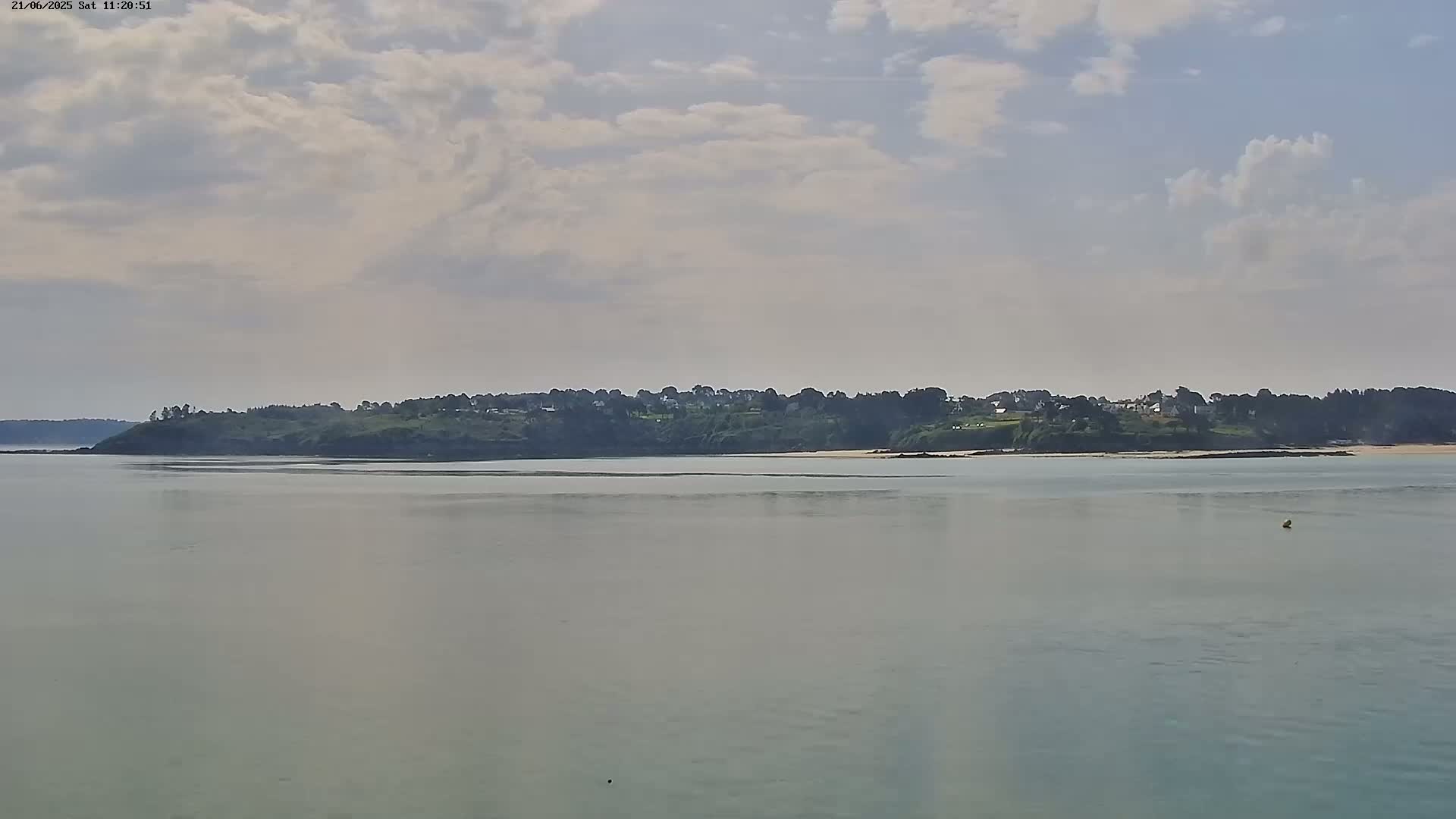 A calm body of water stretches out before a low-lying landmass dotted with houses and trees under a partly cloudy sky.
