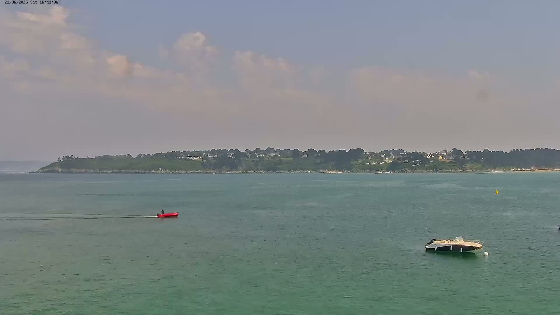A calm, partly cloudy day shows a tranquil body of water with a small red motorboat and a larger, cream-colored boat, and a distant coastline with buildings visible.