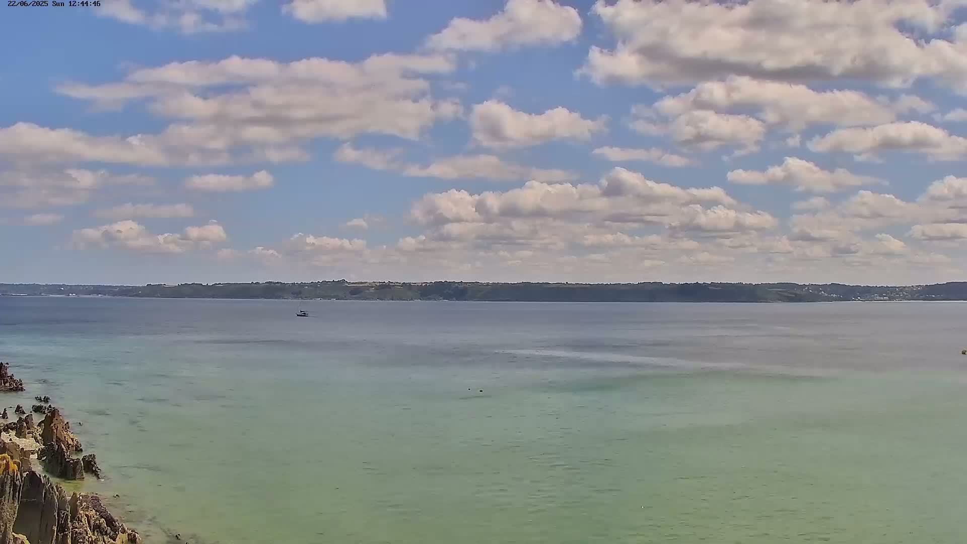 A partly cloudy sky overlooks a calm body of water with a distant shoreline and rocky coast in the foreground.