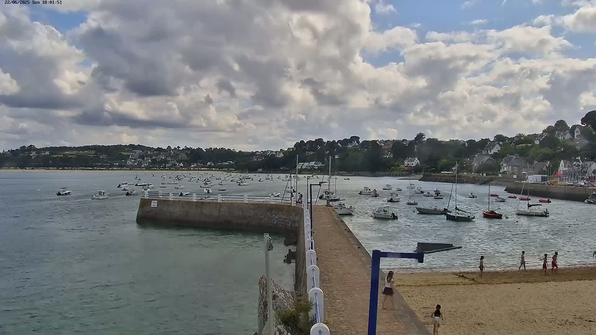 A partly cloudy day shows a sandy beach with several people, a stone pier extending into a harbor filled with numerous boats, and a town nestled on a hillside in the background.