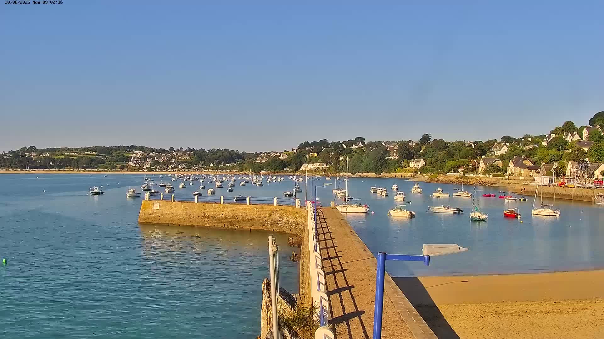 A calm, sunny day shows a harbor filled with numerous boats and small sailboats, nestled next to a shoreline with houses and trees.