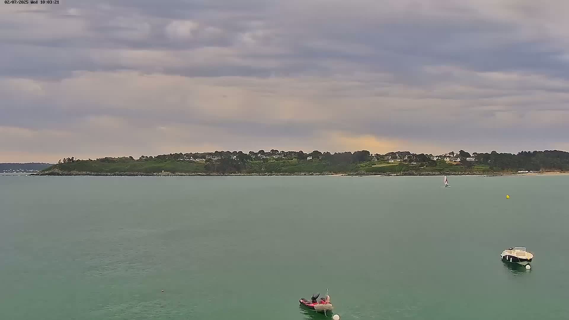 A calm, green body of water with a few small boats, under a mostly cloudy sky, shows a distant shoreline with houses and trees.