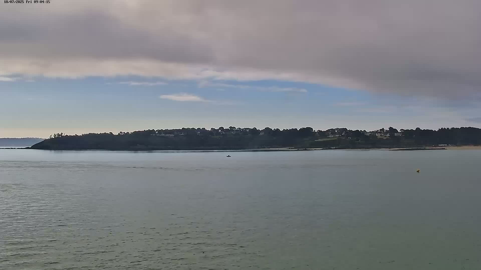 A calm body of water stretches out to a tree-lined shore under a mostly cloudy sky.