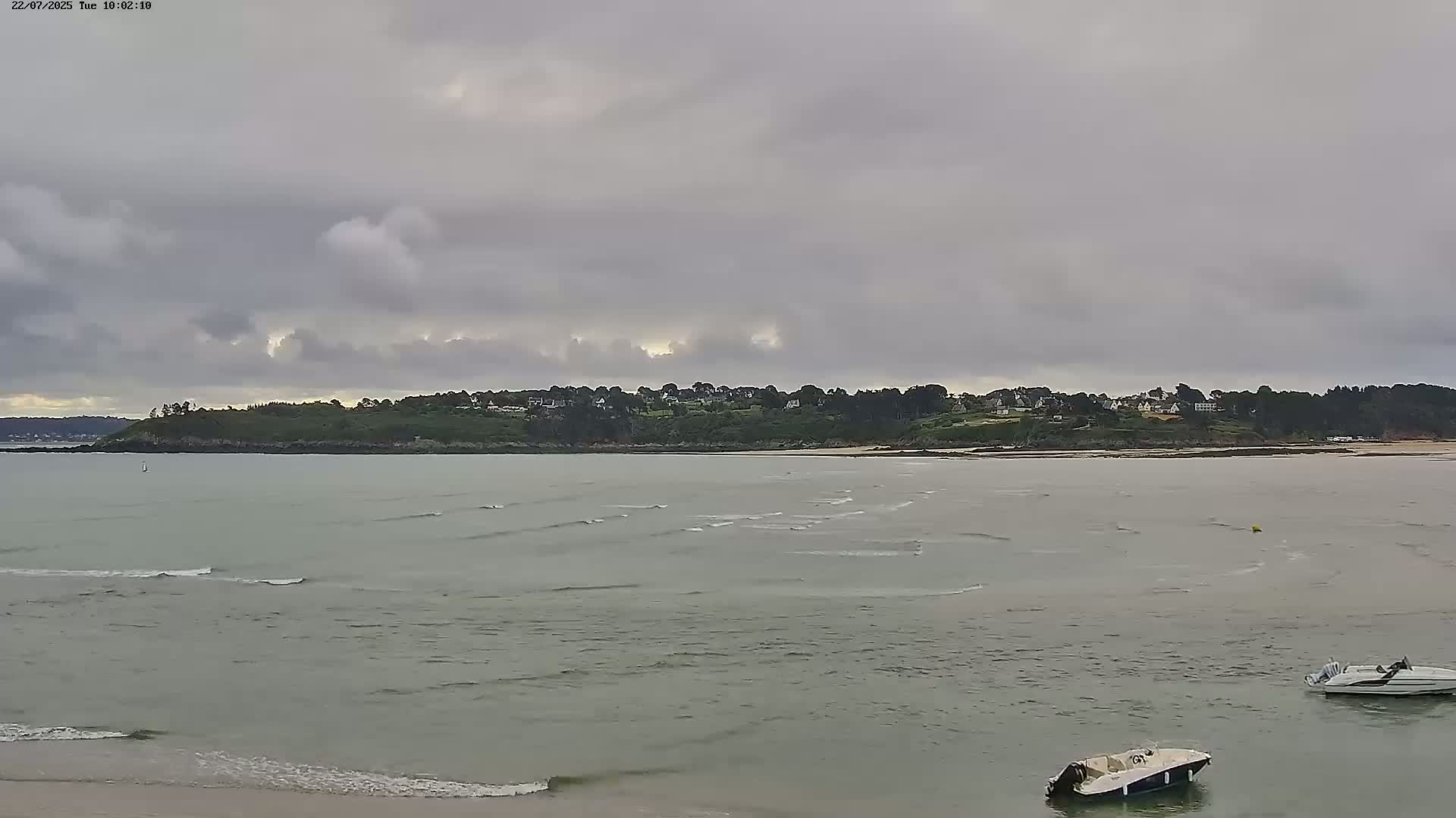 A calm, overcast day at a coastal town, with small waves lapping the shore and two small boats moored in the water.