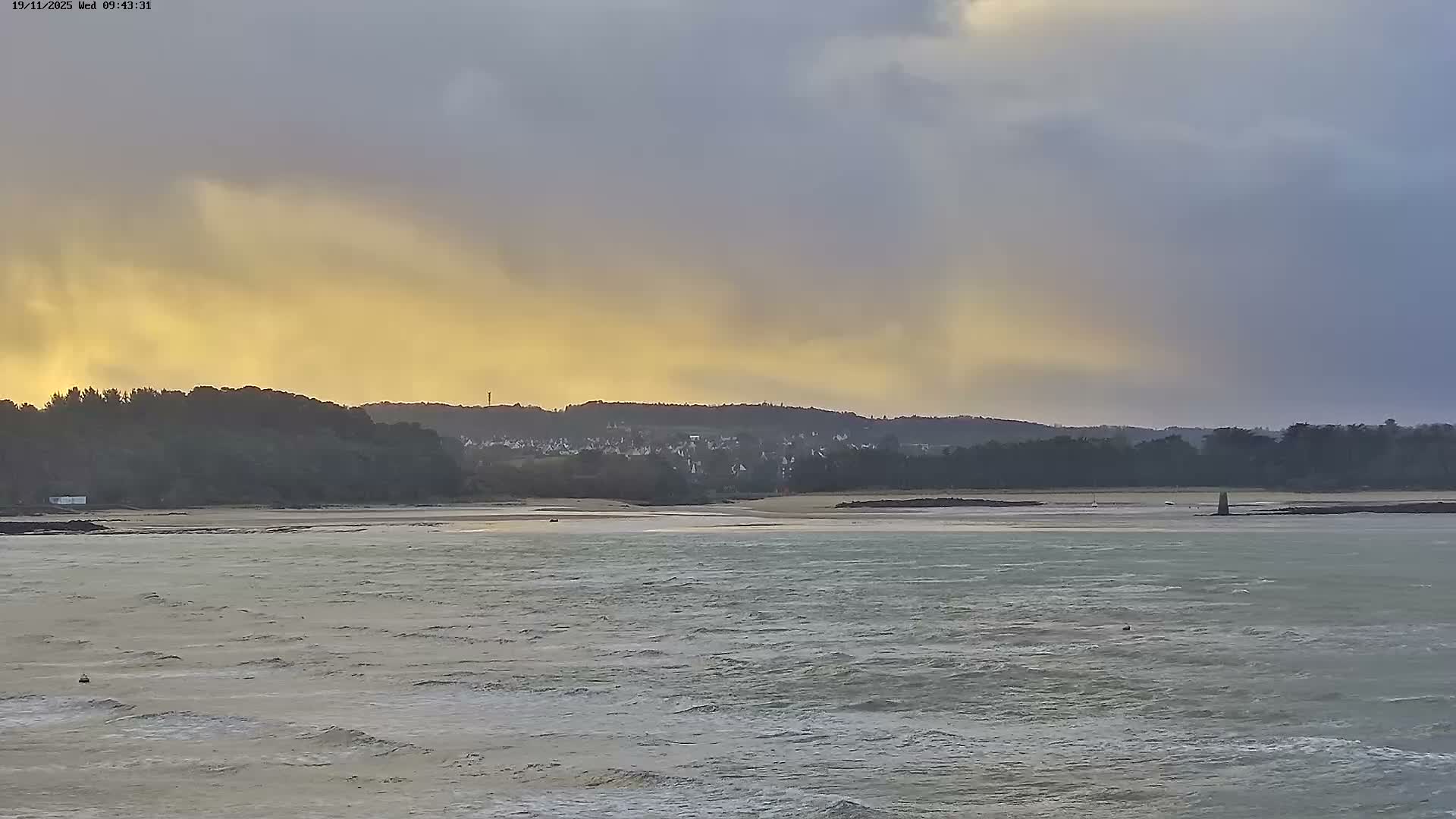 A choppy, grey-green body of water and a sandy shore foreground a distant tree-covered hill and village, all under a dramatic, heavily clouded sky that shifts from dark grey to a vibrant golden-yellow, suggesting overcast conditions with a momentary burst of sunlight.